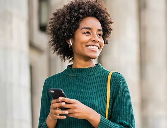 A woman holding her phone with both hands, smiling and looking off into the distance, appears satisfied and relieved, likely pondering, "It was easy to sell my home fast."