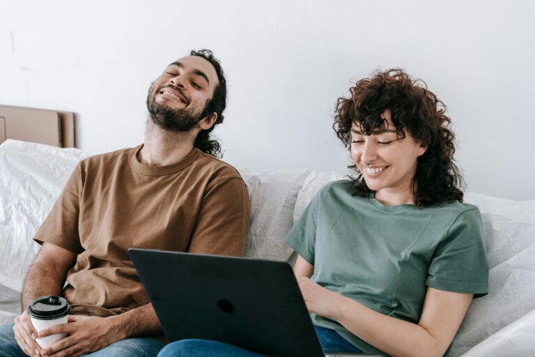 A happy couple is sitting together on a couch. They are smiling and appear relaxed, enjoying each other's company in a cozy living room setting. The background features soft lighting and comfortable home decor, contributing to a warm and inviting atmosphere.