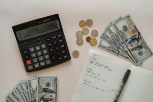 A black calculator beside coins and a notebook on a wooden surface, representing financial planning and budgeting challenges faced by landlords due to a nationwide rent cap.