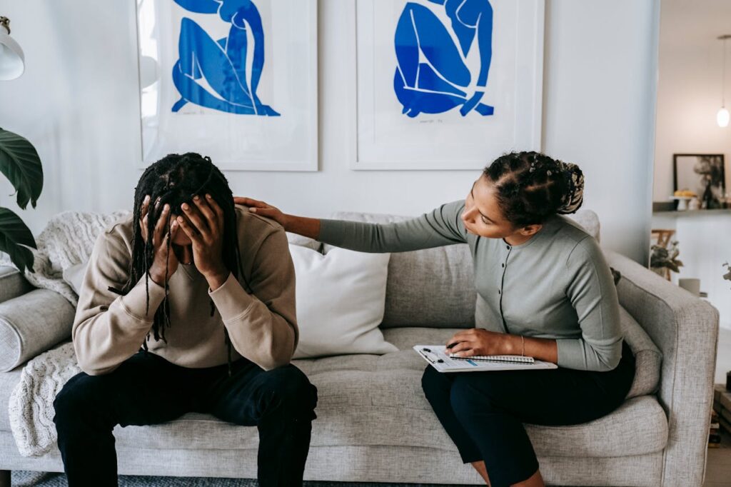 A Black woman consoling an anonymous unhappy boyfriend on a sofa at home, providing emotional support during their efforts to avoid foreclosure.