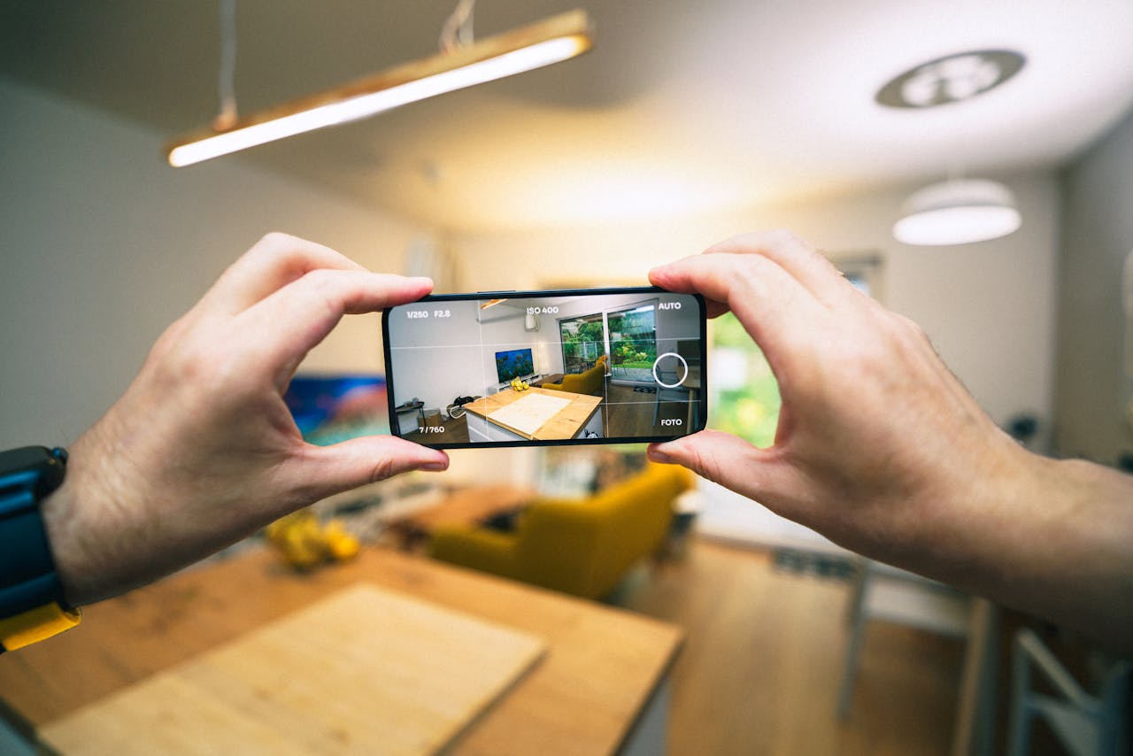 Hands holding a smartphone taking pictures of an apartment to sell a home, featuring a modern living room with bright lighting and contemporary design.